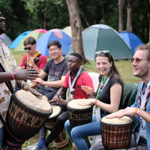 Scouts learn to play drums in front of a backdrop of tents in a forest